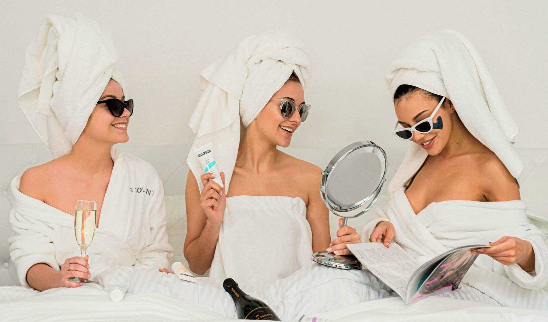 three women in white dressing gowns and white towels on head enjoying champagne